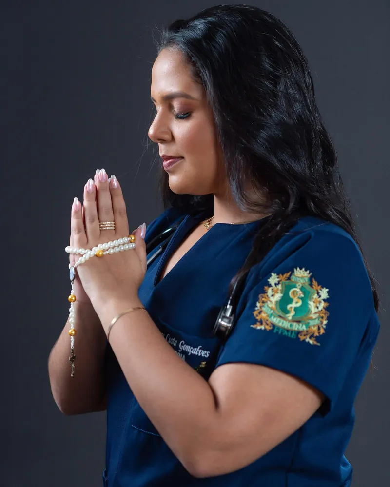 Dra. Lara Costa de scrub azul, de olhos fechados, com as mãos unidas em oração segurando um terço