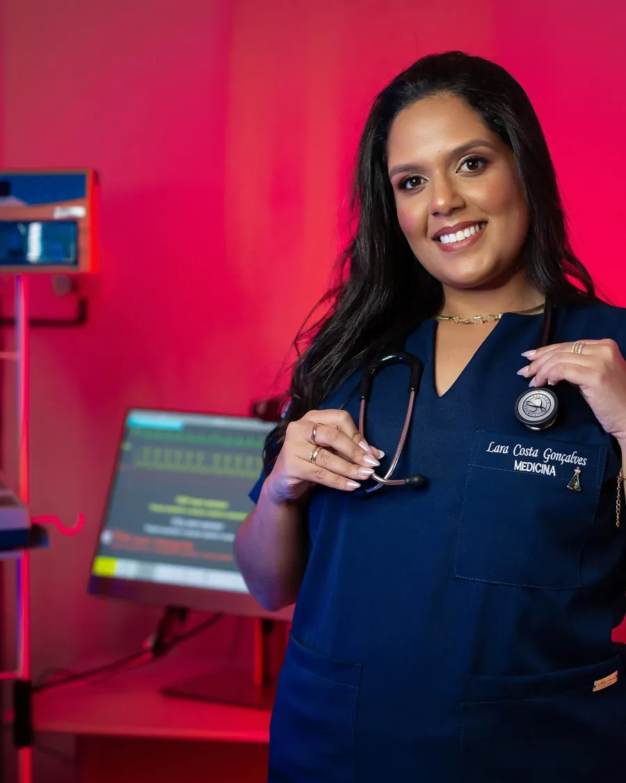 Dra. Lara Costa de scrub azul com estetoscópio, em sala hospitalar com monitor